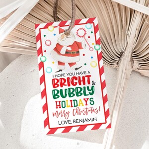 Puede incluir: Una etiqueta de regalo rectangular con un borde a rayas rojas y blancas. La etiqueta presenta un dibujo de Papá Noel sosteniendo una varita mágica. El texto en la etiqueta dice "I HOPE YOU HAVE A BRIGHT & BUBBLY HOLIDAYS merry Christmas! LOVE, BENJAMIN."