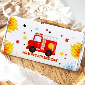 Puede incluir: Un envoltorio de chocolate blanco con un camión de bomberos rojo, llamas amarillas y estrellas negras. El envoltorio dice "Wilson's 5th Birthday".