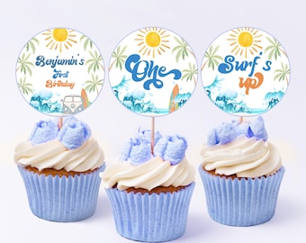 May include: Three blue and white cupcakes with round toppers. The toppers feature a sun, palm trees, and waves. The text on the toppers reads "Benjamin's First Birthday", "One", and "Surf's Up".