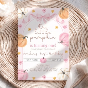 May include: A pink and white invitation card with watercolor pumpkins and floral accents. The text reads "Our little pumpkin is turning one!" and includes party details. The card is set on a woven, beige surface.