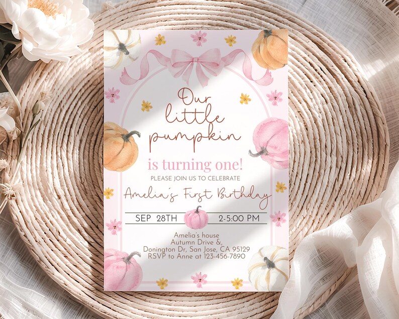 May include: A pink and white invitation card with watercolor pumpkins and floral accents. The text reads "Our little pumpkin is turning one!" and includes party details. The card is set on a woven, beige surface.