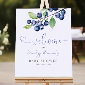 Op de afbeelding: Een wit welkomsbord met een aquarel-illustratie van blauwe bessen aan een tak. Het bord zegt "Welkom bij Emily Brown's Baby Shower op 28 juli 2058".