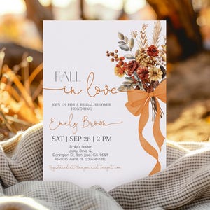 May include: A bridal shower invitation with the text "Fall in Love" and details for Emily Brook's shower. The invitation features a floral bouquet with orange ribbon and is set against a blurred background.