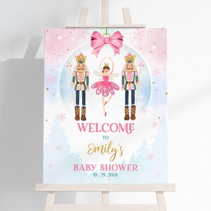 Puede incluir: Un cartel de bienvenida para un baby shower con un fondo de acuarela rosa, azul y blanco. El cartel presenta una bailarina y dos figuras de cascanueces dentro de una bola de nieve. El texto dice "Bienvenido a la fiesta de bienvenida de Emily 10.25.2058".