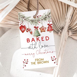 Op de afbeelding: Een rechthoekig cadeaulabel met een bruin touwtje, met de tekst "BAKED with love merry Christmas FROM THE BROWN" in rood en zwart. Het label is versierd met aquarel illustraties van kerstkoekjes en groen.