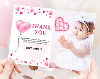 May include: Pink thank you card with a pink heart balloon and a photo of a baby girl wearing a white dress and a white headband. The text on the card reads "Thank You" and "I enjoyed the cake and the presents too, but my favorite part was seeing you! Thank you for coming to my party! Love, Amelia."