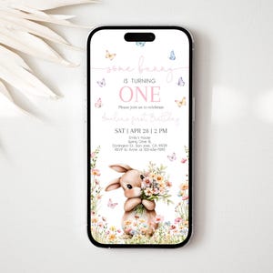 Może przedstawiać: Smartfon wyświetla cyfrowe zaproszenie na pierwsze urodziny. Zaproszenie zawiera ilustrację królika z kwiatami i tekst "some bunny is turning ONE". Motyle i elementy kwiatowe zdobią projekt.