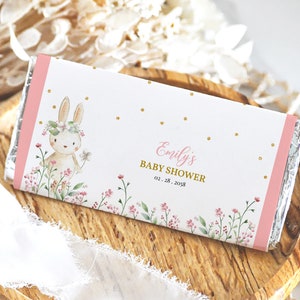 May include: A pink and white chocolate bar wrapper with a gold foil accent. The wrapper features a watercolour illustration of a bunny wearing a flower crown and holding a flower. The text on the wrapper reads "Emily's Baby Shower 02.28.2028".