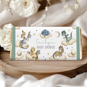 May include: A baby shower chocolate bar wrapper with watercolour dragons on clouds, stars, and a crescent moon. The wrapper has a light blue and white background with the text "Emily's BABY SHOWER". The ends of the wrapper are light green.