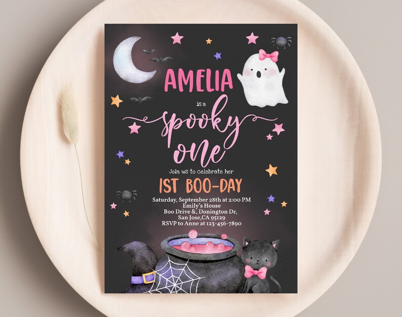 May include: A black and white invitation card with a pink and white ghost, a cauldron, and a black cat. The text on the card reads "Amelia is a Spooky One" and "1st Boo-Day".