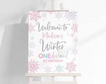 May include: Welcome to Madison's Winter Wonderland 1st Birthday sign with a white background and snowflakes in pink, purple, and blue. The text is in silver glitter.