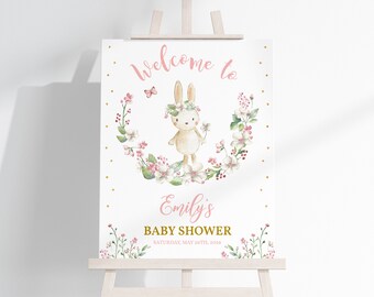 Könnte beinhalten: Willkommen zur Babyparty von Emily Schild mit einem weißen Kaninchen, das eine rosa Blumenkrone trägt und eine Blume hält. Das Kaninchen ist von einem Kranz aus rosa Blumen und grünen Blättern umgeben. Das Schild ist mit goldenen Punkten verziert.