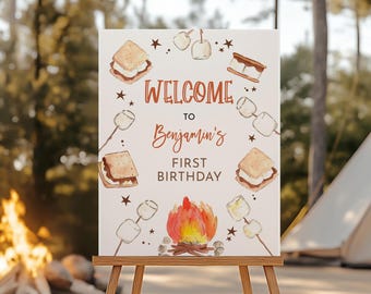 May include: A sign for Benjamin's first birthday, with a campfire theme. The sign features illustrations of s'mores, marshmallows, and a campfire. The text reads "Welcome to Benjamin's First Birthday."