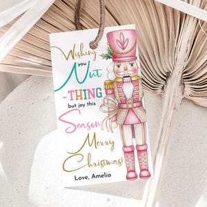 May include: A white gift tag with a watercolor illustration of a pink nutcracker soldier. The tag has the text "Wishing you Nut-THING but joy this Season Merry Christmas! Love, Amelia."