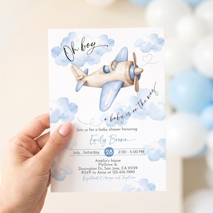 May include: Baby shower invitation featuring a watercolor illustration of a light blue airplane with a propeller, surrounded by blue clouds. The text "Oh boy" and "a baby is on the way" are included.