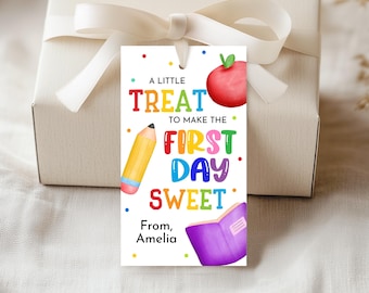 Könnte beinhalten: Ein Geschenkanhänger mit dem Text "A LITTLE TREAT TO MAKE THE FIRST DAY SWEET" mit einem roten Apfel, einem gelben Bleistift und einem lila Buch. Der Anhänger ist an einer weißen Geschenkbox mit einem weißen Band befestigt.