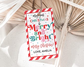 May include: A rectangular Christmas gift tag with a red and white striped border. The tag features the text "May your CHRISTMAS be Merry and Bright" in various colors, along with "merry Christmas!" and "LOVE, AMELIA". A brown string is attached.