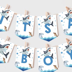 Puede incluir: Una pancarta de baby shower azul y blanca con las palabras "It's a Boy" en letras azules. Cada letra está decorada con un tiburón de acuarela y burbujas.
