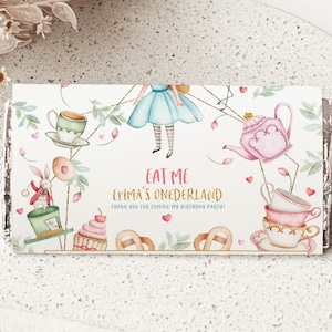 Puede incluir: Envoltorio de barra de chocolate con temática de "Alicia en el país de las maravillas". El diseño presenta ilustraciones de Alicia, tazas de té, una tetera y un conejo. El texto incluye "EAT ME" y "Emma's Onerderland".