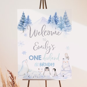 May include: A blue and white winter wonderland themed first birthday party sign with the text "Welcome to Emily's One derland 1st Birthday". The sign features a snowy mountain, pine trees, snowflakes, and a polar bear, penguin, seal, and owl.
