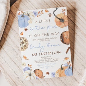 Puede incluir: Invitaci&oacute;n a un baby shower con calabazas de acuarela, tarta y detalles florales. El texto dice "A Little Cutie Pie Is On The Way" con el nombre de la homenajeada, la fecha, la hora y el lugar. Sobre una superficie de madera.