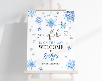 Puede incluir: Un letrero blanco sobre un caballete de madera con copos de nieve azules de acuarela y texto. El texto dice: "Un pequeño copo de nieve está en camino, bienvenido al baby shower de Emily."