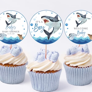 May include: Three blue and white cupcake toppers featuring a shark, dolphin, and bubbles. The toppers read "Emily's Baby Shower", "It's a Boy", and "Welcome Baby".