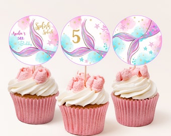 May include: Three pink cupcakes with white frosting and mermaid tail cupcake toppers. The toppers are round and feature a watercolor background with a mermaid tail, stars, and gold glitter. One topper says "Splish Splash", another says "Amelia's 5th Birthday", and the third has the number "5" on it.