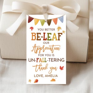 May include: A white gift tag with a fall-themed design. The tag features the text "YOU BETTER BE-LEAF OUR Appreciation FOR YOU IS UN-FALL-TERING thank you LOVE, AMELIA" with colorful leaf and banner illustrations. The tag is tied to a gift box with a white ribbon.