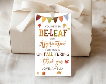 May include: A white gift tag with a fall-themed design. The tag features the text "YOU BETTER BE-LEAF OUR Appreciation FOR YOU IS UN-FALL-TERING thank you LOVE, AMELIA" with colorful leaf and banner illustrations. The tag is tied to a gift box with a white ribbon.