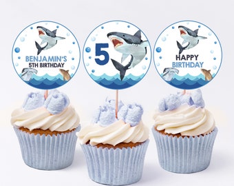 May include: Three blue and white cupcake toppers with a shark design. The toppers feature a shark with a white background and blue waves. The text on the toppers reads "Benjamin's 5th Birthday", "Happy Birthday", and "5".