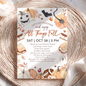 Op de afbeelding: Een uitnodiging met een herfstthema, met illustraties van pompoenen, een vleermuis en s'mores. De tekst luidt "Let's Get Together and Enjoy All Things Fall" met evenementdetails voor 28 oktober om 14.00 uur. De uitnodiging ligt op een gevlochten placemat.