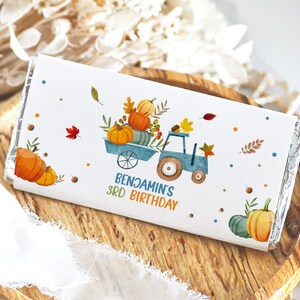 May include: A white chocolate bar wrapper with a fall-themed design. A blue tractor is hauling a load of pumpkins. The wrapper says "Benjamin's 3rd Birthday".
