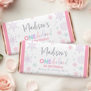 Könnte beinhalten: Zwei Schokoriegelverpackungen mit Winterwunderland-Motiv. Die Verpackungen sind weiß mit Schneeflockenmustern in Pastellfarben und dem Text "Madison's ONEderland 1st birthday". Rosa und goldfarbene Akzente vervollständigen das Design.