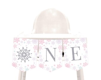 May include: A high chair decorated with a banner for a first birthday. The banner is white with snowflake patterns in pink, purple, and blue. The word "ONE" is spelled out in silver glitter letters.