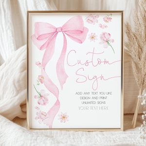 May include: A framed print with a gold border features a watercolor design. A large pink bow and flowing ribbons are at the top, with floral accents. The text "Custom Sign" is displayed, along with additional text below.