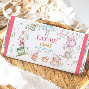 May include: A white chocolate bar wrapper with a pink border. The wrapper features an illustration of Alice in Wonderland characters and the text "Eat Me Emma's Baby Shower 05.25.2058".