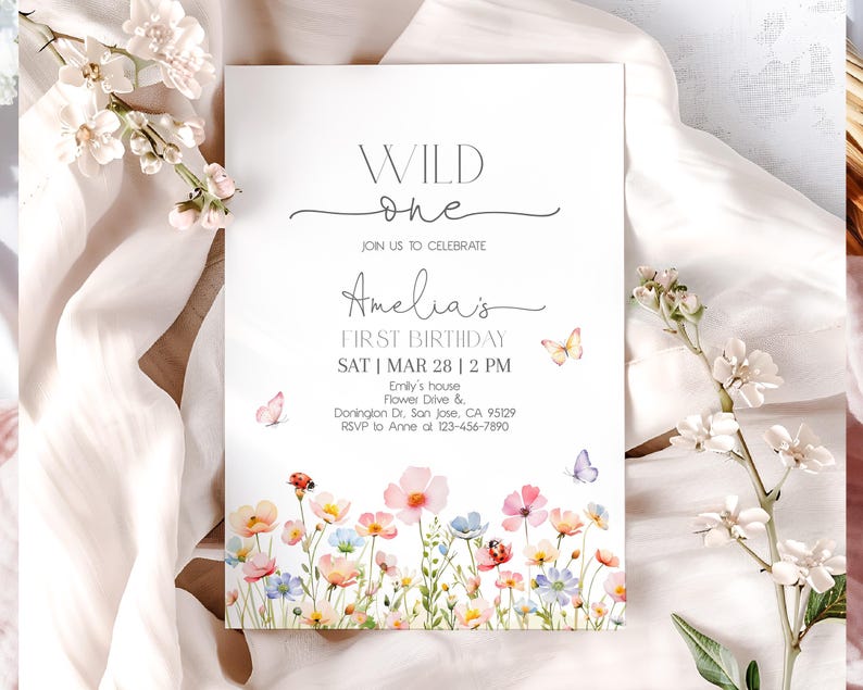 以下が含まれることがあります： 「WILD one」と「Amelia's FIRST BIRTHDAY」の文字が入った白い招待状。水彩画のフローラルボーダーと蝶が特徴です。招待状には、イベントの日時と場所が記載されています。