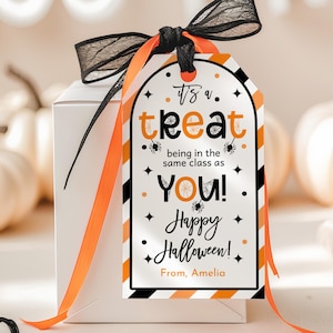 Puede incluir: Una caja de regalo blanca con una etiqueta temática de Halloween. La etiqueta tiene rayas naranjas y negras, las palabras "It's a treat" y "Happy Halloween!" con gráficos de arañas y estrellas. Está atada con una cinta negra y naranja.