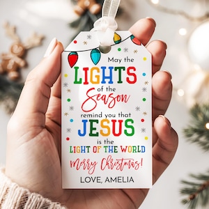 Könnte beinhalten: Ein weißes Weihnachtsgeschenkanhänger mit einem Band, gehalten in einer Hand. Das Etikett enthält farbigen Text mit der Aufschrift "May the LIGHTS of the Season remind you that JESUS is the LIGHT OF THE WORLD merry Christmas! LOVE, AMELIA."