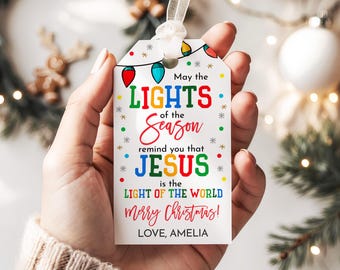 May include: A white Christmas gift tag with a ribbon, held in a hand. The tag features colorful text that reads "May the LIGHTS of the Season remind you that JESUS is the LIGHT OF THE WORLD merry Christmas! LOVE, AMELIA."