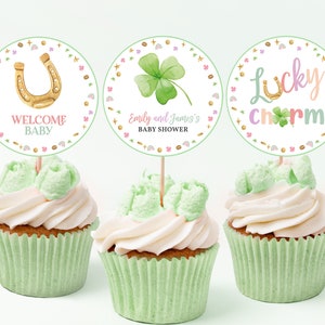 May include: Three green cupcake wrappers with white frosting and gold cupcake toppers. The toppers feature a horseshoe, a four-leaf clover, and the text "Lucky Charm".