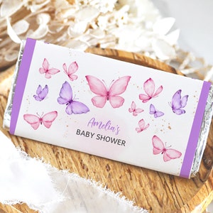Puede incluir: Un envoltorio de chocolate blanco con un diseño de mariposas moradas y rosas. El envoltorio dice "Baby Shower de Amelia" en letras doradas.