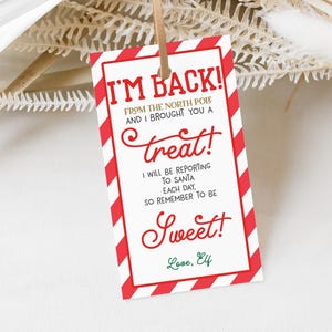 Puede incluir: Una etiqueta de regalo rectangular con un borde a rayas rojas y blancas y el texto "I'M BACK!" en rojo. La etiqueta incluye las frases "FROM THE NORTH POLE AND I BROUGHT YOU A treat!" y "SO REMEMBER TO BE Sweet!" con la firma "Love, Elf."