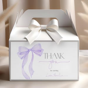 Puede incluir: Una caja de regalo blanca con asa y lazo blanco en la parte superior. La parte delantera de la caja presenta un lazo lavanda estilo acuarela y las palabras "THANK you for coming Love, Amelia."