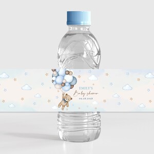 Może przedstawiać: Etykieta na butelkę wody na baby shower w kolorze niebieskim i brązowym z pluszowym misiem trzymającym balony, chmury i gwiazdki. Tekst na etykiecie brzmi "Emily's Baby Shower 09.28.2018".