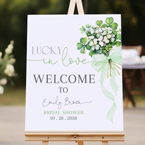 May include: A white welcome sign on a wooden easel with the text "Lucky in Love Welcome to Emily Brown Bridal Shower 10.28.2058". The sign features a bouquet of white flowers and green shamrocks with a green ribbon.