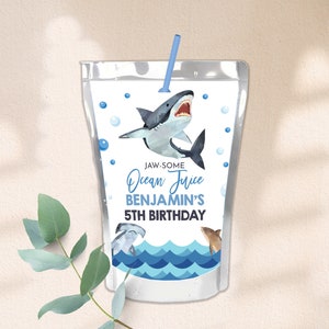 May include: A blue and white juice box with a shark design. The text on the box reads "Jaw-some Ocean Juice Benjamin's 5th Birthday".