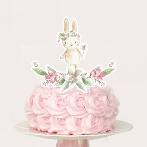 May include: A pink frosted cake with a white bunny wearing a flower crown and holding a flower. The bunny is surrounded by green leaves and pink flowers.