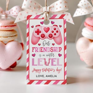 May include: A Valentine's Day gift tag with a pink video game controller graphic and the text "Our Friendship is on another LEVEL". The tag is decorated with hearts and diagonal stripes. A clear ornament with macarons and a heart-shaped candy is in the background.
